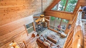A wooden cabin interior with a loft bedroom, a fireplace, a sofa, and large windows offering a view of the outdoors.