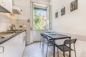 A compact kitchen with a gas stove, a kettle, and a window overlooking a building. There is a dining area with a table and chairs.