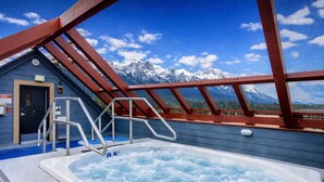 A hot tub with a view of snow-capped mountains, a wooden deck, and a blue building with a door. - Heart of Canmore - Pet Friendly with Shared Rooftop Hot Tub (Canmore)