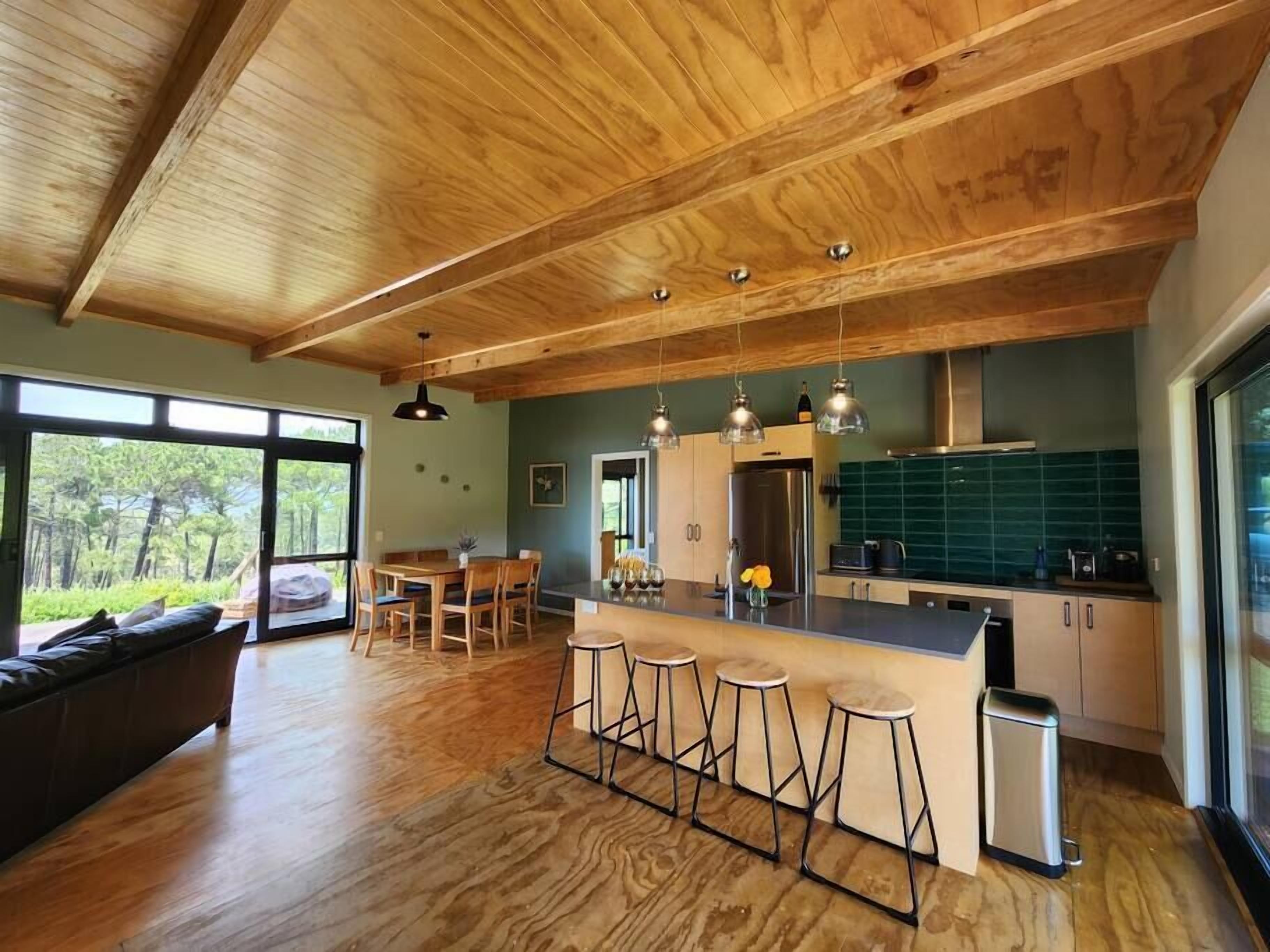 A modern kitchen with wooden ceiling beams, a central island with bar stools, a dining area with a table and chairs, and large windows offering a view of the outdoors.