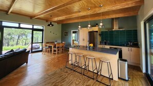 A modern kitchen with wooden ceiling beams, a central island with bar stools, a dining area with a table and chairs, and large windows offering a view of the outdoors. - Hahei Hideout Retreat - private and surrounded by bush. Pond and small orchard (Hahei)