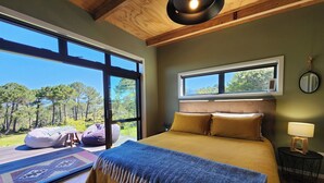 A bedroom with a large window, a bed with a blue throw, and a view of a forest. - Hahei Hideout Retreat - private and surrounded by bush. Pond and small orchard (Hahei)