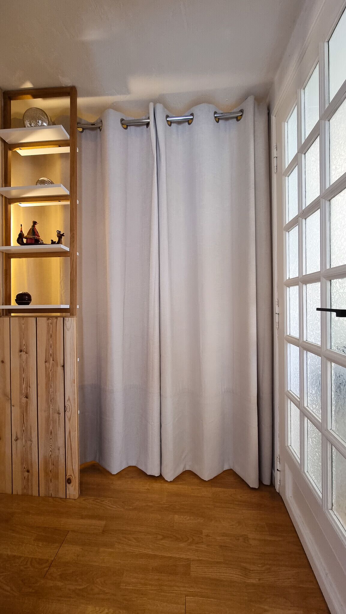 A room with wooden flooring, a white curtain, a shelf with decorative items, and a window with frosted glass.