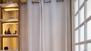 A room with wooden flooring, a white curtain, a shelf with decorative items, and a window with frosted glass.