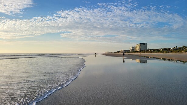 Een strand met een rustige zee, een paar mensen die wandelen, en een gebouw op de achtergrond.