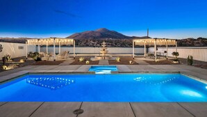 Piscina con montañas al fondo, mobiliario exterior y una pérgola con luces colgantes.