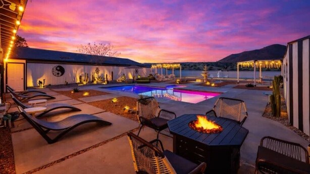 Zona de piscina con sillas de descanso y un brasero, rodeada de edificios y un cielo al atardecer.