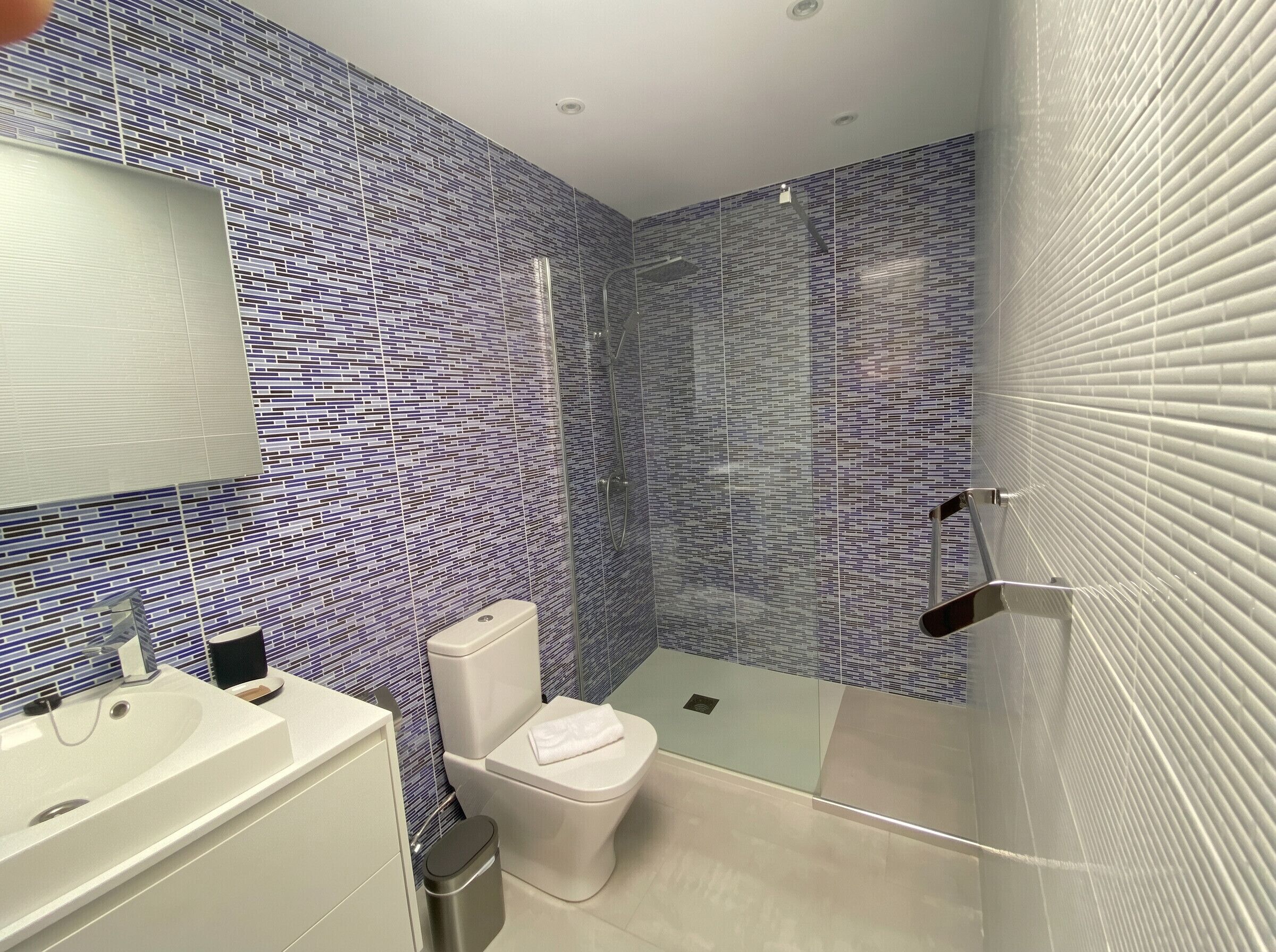 Un baño moderno con pared de azulejos estampados, cabina de ducha de vidrio, inodoro blanco y lavamanos con espejo arriba.