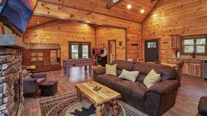 A cozy log cabin interior with a living room, a stone wall, a fireplace, and a kitchen area.
