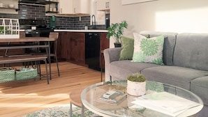 Una sala moderna con un sofá gris, una mesa de centro de vidrio y una mesa de comedor de madera. Se observa una cocina con armarios blancos y una estufa negra. Una ventana muestra vistas a árboles.