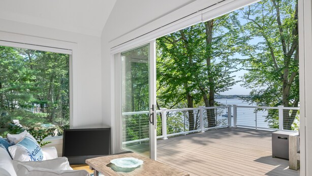 A modern living room with a large sliding glass door leading to a wooden deck with a view of a lake and trees. - Luxurious island home on the water, 30 minute ferry ride from Portland Maine (Portland)