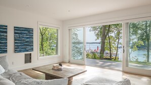 A modern living room with a large sliding glass door leading to a balcony with a view of the water and trees. - Luxurious island home on the water, 30 minute ferry ride from Portland Maine (Portland)