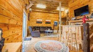 A wooden cabin interior with a leather sofa, a round rug, a bar area with stools, and a mounted television. - Cozy mountain retreat with spectacular view to unwind and relax. Pet friendly ! (Glade Valley)