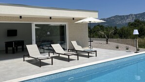A modern outdoor pool area with lounge chairs and a table, a clear blue sky, and mountains in the background.
