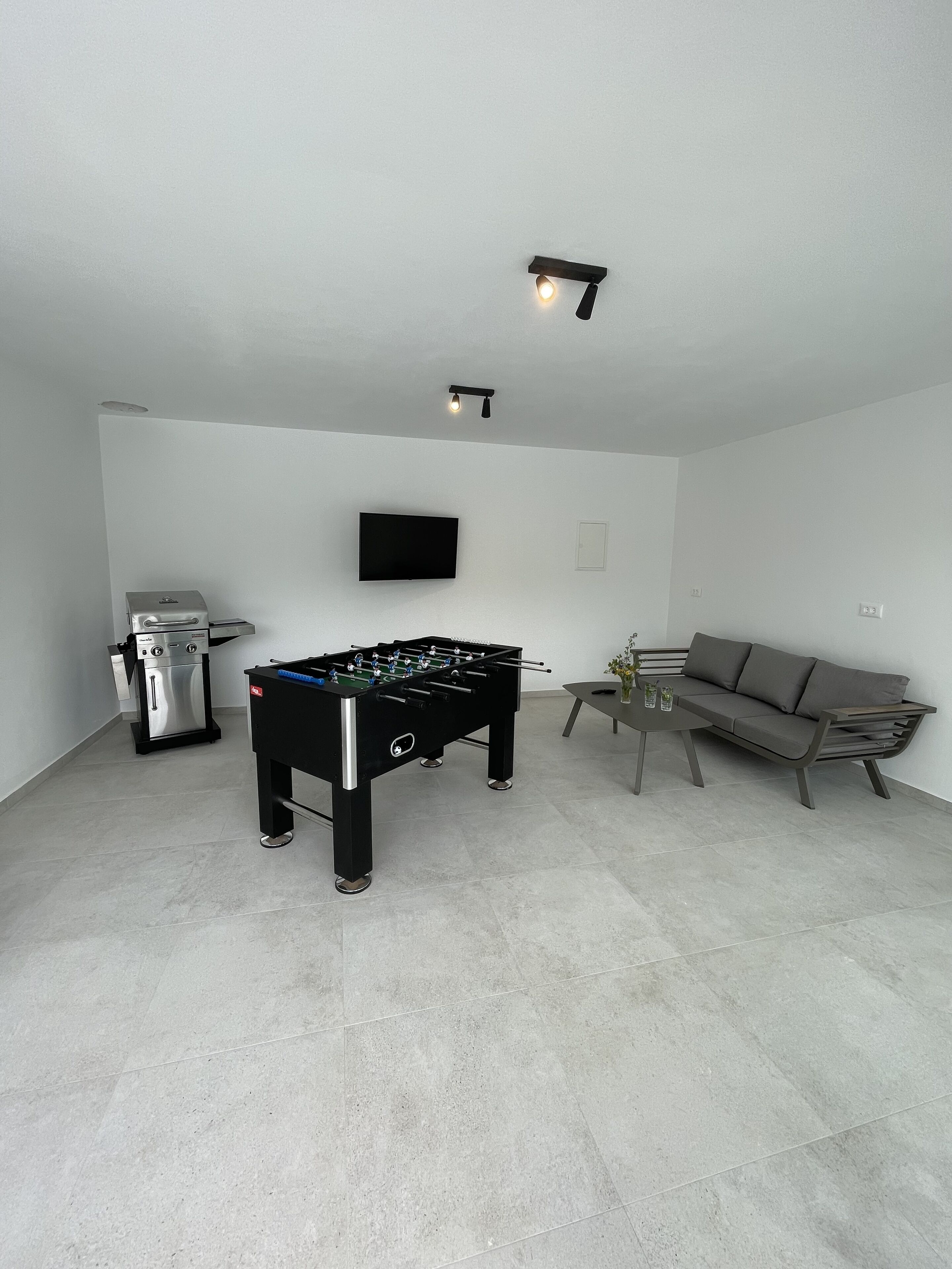 Un salon moderne comprenant une table de baby-foot, un canapé, une table basse, un téléviseur fixé au mur et un petit coin cuisine avec une machine à café.
