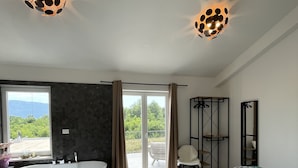 A modern bedroom with a large bed, a freestanding bathtub, and a balcony with a view of greenery.