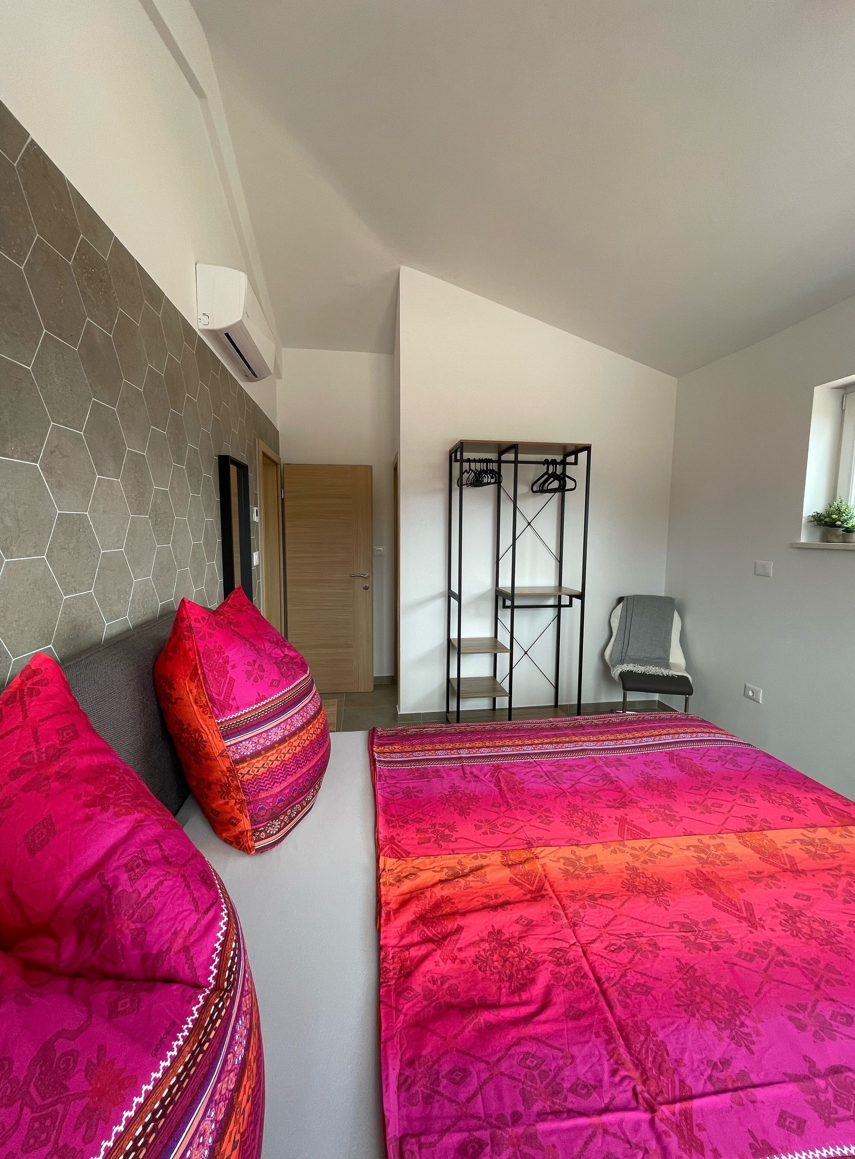 Une chambre à coucher avec un lit, deux coussins colorés, une armoire et un portant à vêtements.