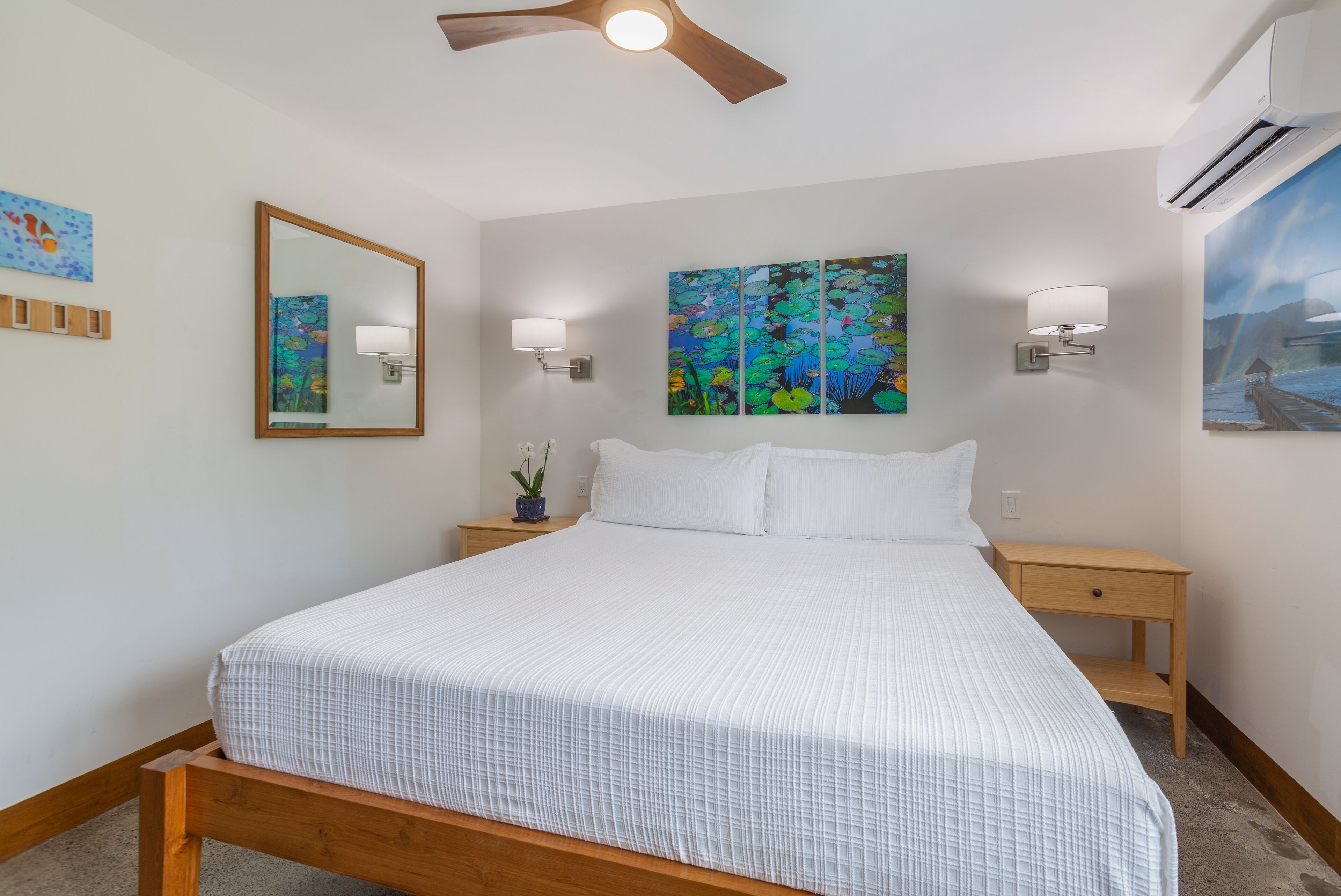 Une chambre à coucher avec un lit en bois, du linge de lit blanc, un ventilateur de plafond, des lampes fixées au mur, un miroir et des œuvres d’art.
