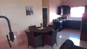 A compact living space with a dining area featuring a round table and wicker chairs, a kitchenette with dark cabinets, and a wall-mounted picture of a cityscape. - Loodik Vacation Home Brompton Manor  (Speculation)
