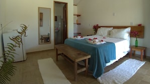 A bedroom with a bed, a bedside table, a bench, a mirror, a sink, and a door leading to a bathroom. - POUSADA PEDRA DO NEGRO (Porto Seguro)