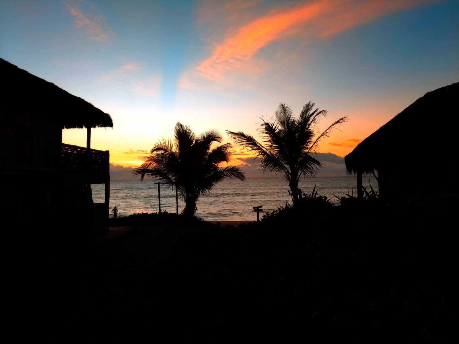 A beach scene with thatched-roof huts and palm trees silhouetted against a sunset.