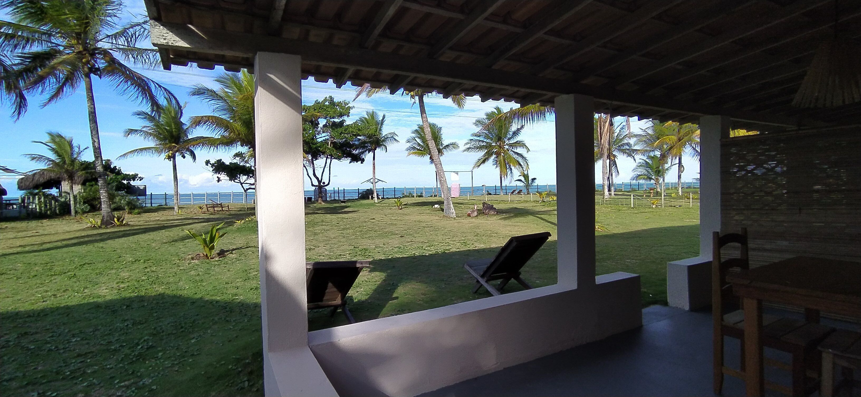 A covered outdoor area with a view of the sea, palm trees, and a grassy lawn.