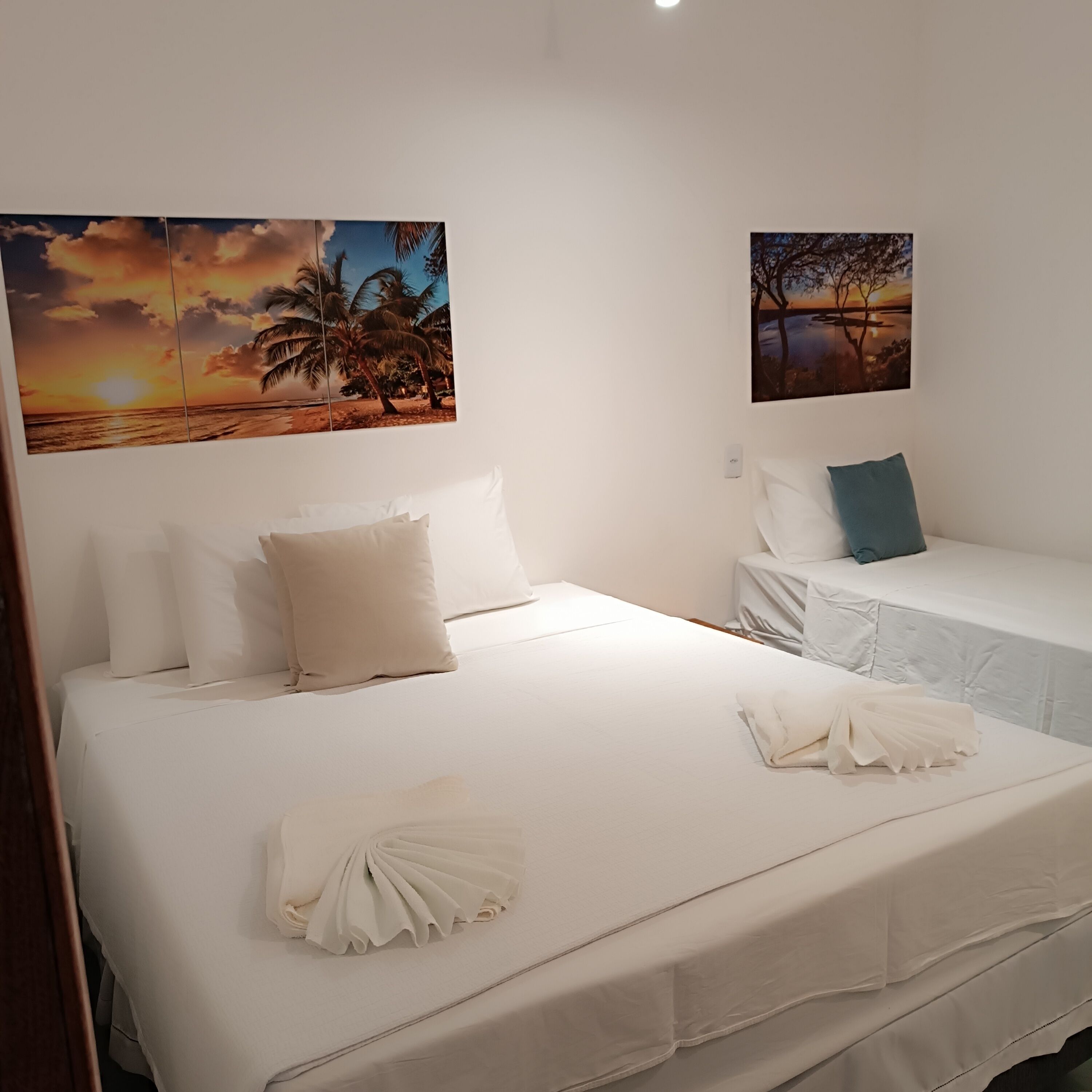 A hotel room with a large bed, two single beds, and two wall-mounted paintings of tropical scenes.
