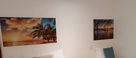 A hotel room with a large bed, two single beds, and two wall-mounted paintings of tropical scenes.
