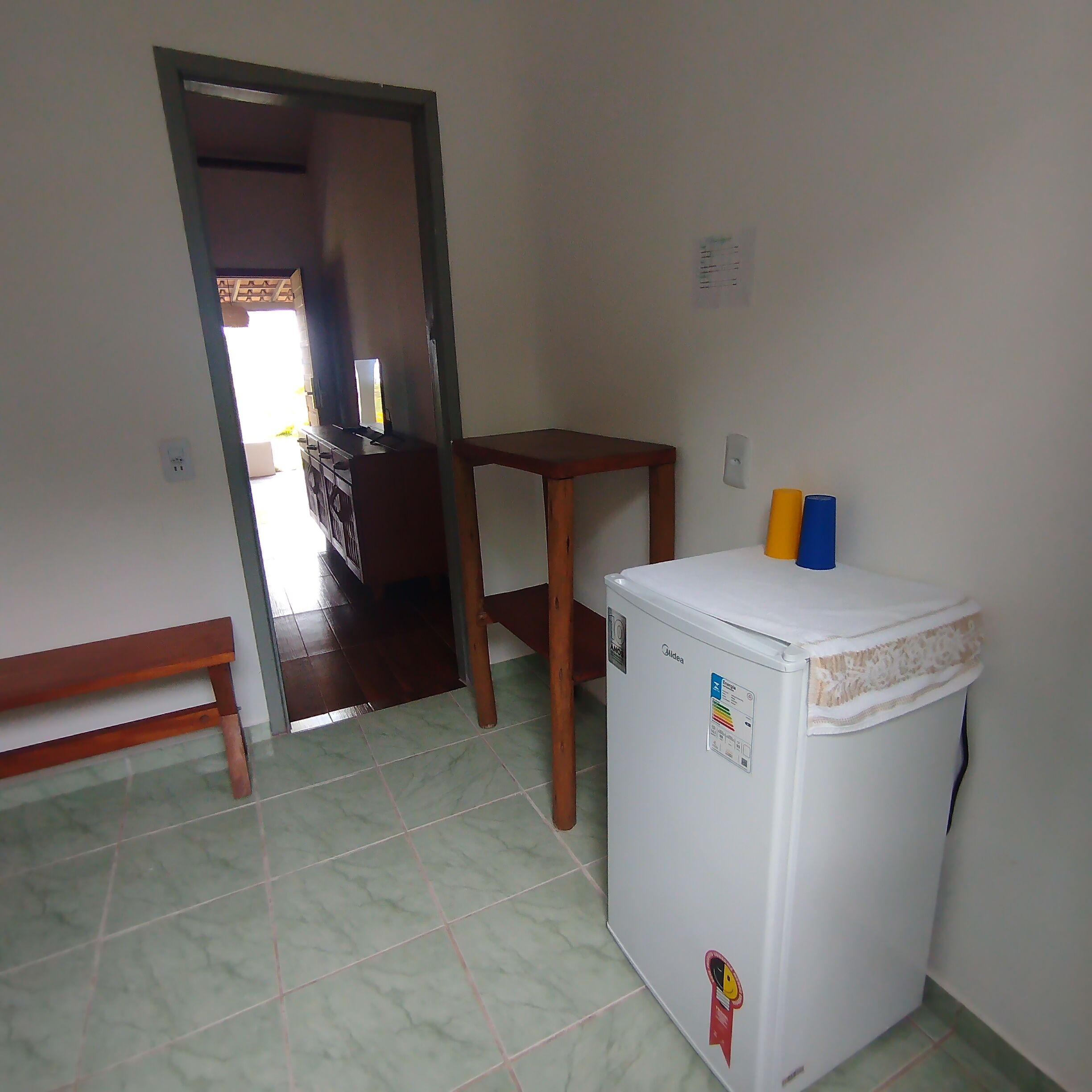 A compact room with a refrigerator, a wooden bench, a small table, and a door leading to another room.