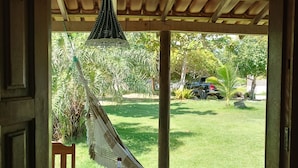 A hammock under a wooden pergola, a wooden table, and a view of a grassy yard with trees and a parked vehicle. - POUSADA PEDRA DO NEGRO (Porto Seguro)