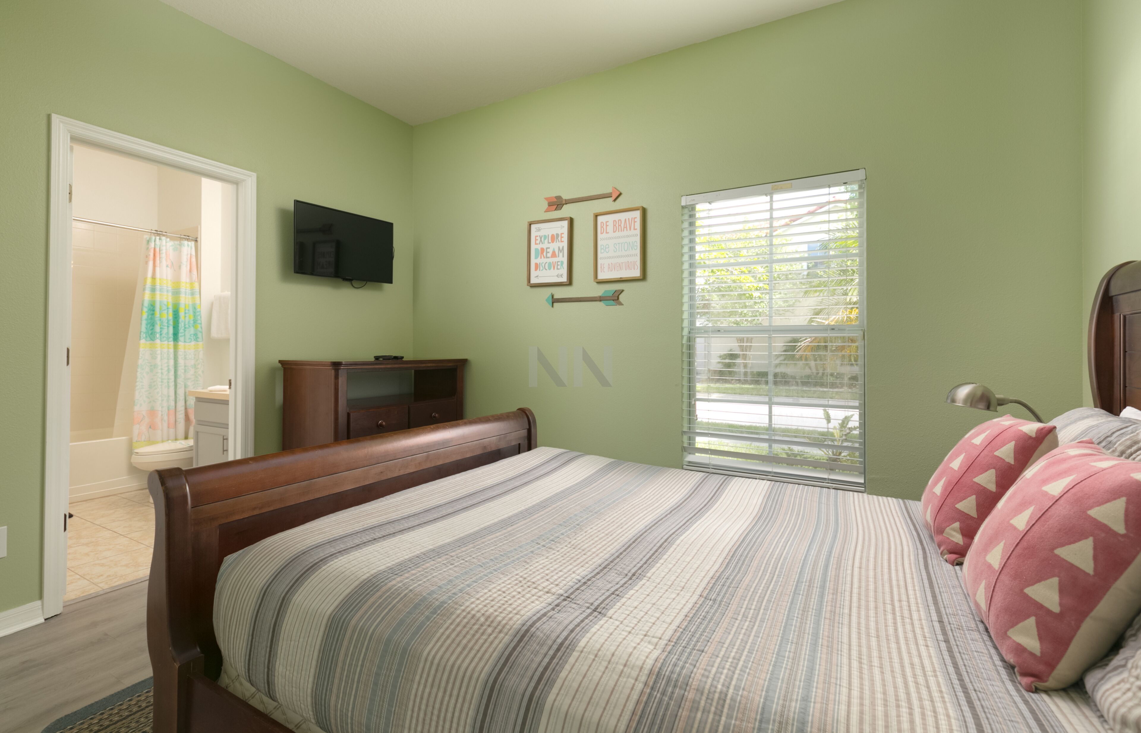 A bedroom with a bed, a television on the wall, a window with blinds, and motivational posters.