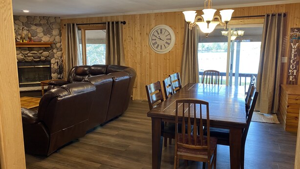 A cozy living room with a leather sofa, a dining table with chairs, a stone fireplace, and a large clock on the wall. - Lakefront Cabin on a Private Lake - only a 0.5 mile from the snowmobile trials! (Crandon)