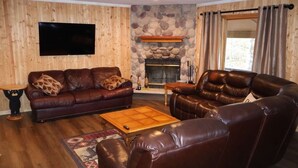 A living room with a stone fireplace, leather sofas, a flat-screen TV, and wooden walls. - Lakefront Cabin on a Private Lake - only a 0.5 mile from the snowmobile trials! (Crandon)