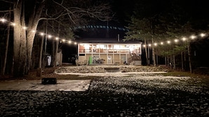 A two-story building with a deck and outdoor seating area, surrounded by trees and decorated with string lights. - Lakefront Cabin on a Private Lake - only a 0.5 mile from the snowmobile trials! (Crandon)