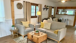Ein gemütliches Wohnzimmer mit grauem Sofa, zwei Sesseln, einem Holztisch und einem gemusterten Teppich. Im Hintergrund ist eine Küche zu sehen.