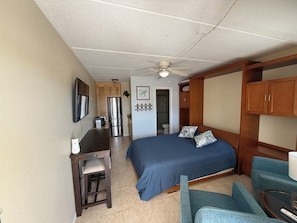 Una habitación compacta con cama, escritorio, silla, ventilador de techo y kitchenette.