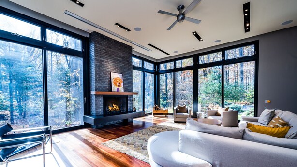 A modern living room with a fireplace, large windows, and a view of the forest. - Harbert Home With Private Pool & Hot Tub! Close To Beach!! (Harbert)