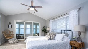 Une chambre à coucher avec un lit, un ventilateur de plafond, une fenêtre avec des stores, une chaise et une vue sur l’extérieur.