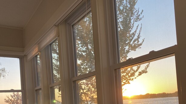 Une pièce dotée d’une grande fenêtre donnant sur une étendue d’eau au coucher du soleil. On y trouve une chaise et un lit avec un oreiller à motifs.