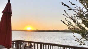 Une terrasse en bois avec une table et des chaises, donnant sur une étendue d’eau au coucher du soleil.