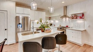 A modern kitchen with white cabinetry, stainless steel appliances, a central island with seating, pendant lighting, and a backsplash with a subtle pattern. - The Lischey House-East Nashville (Nashville)