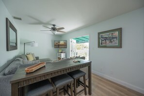 Un coin repas avec une table et des bancs en bois, un ventilateur de plafond, un canapé avec des coussins, une lampe et un tableau encadré au mur.