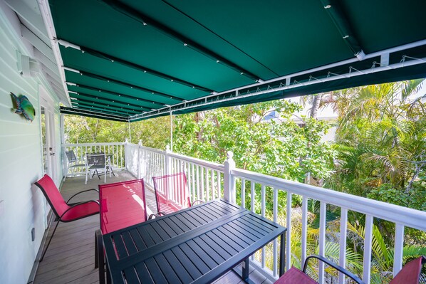 A covered outdoor area with a table and chairs, surrounded by a white railing and lush greenery.