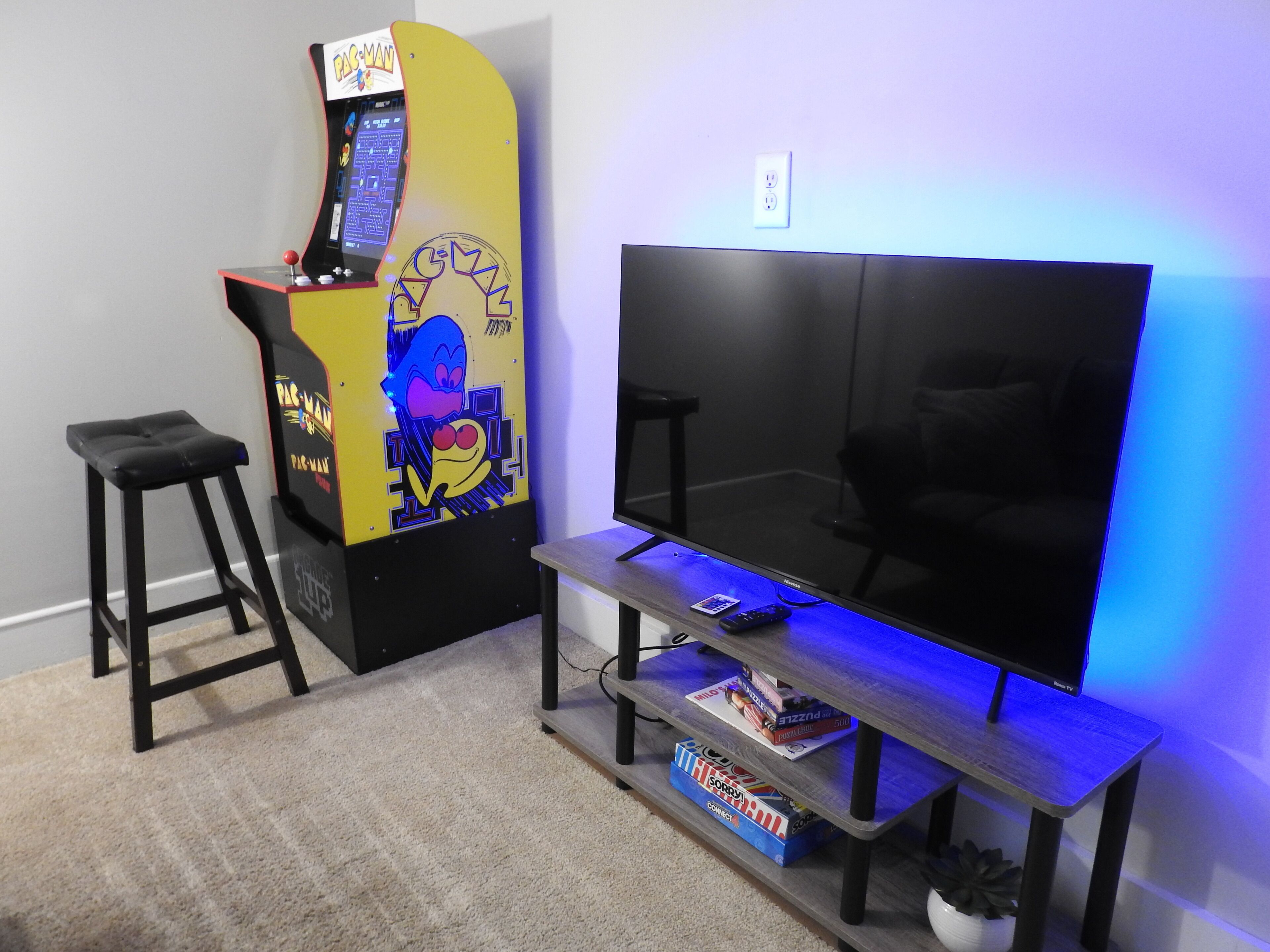 A living room with a large flat-screen TV, a gaming console, a stool, and a small table with books and a remote control.