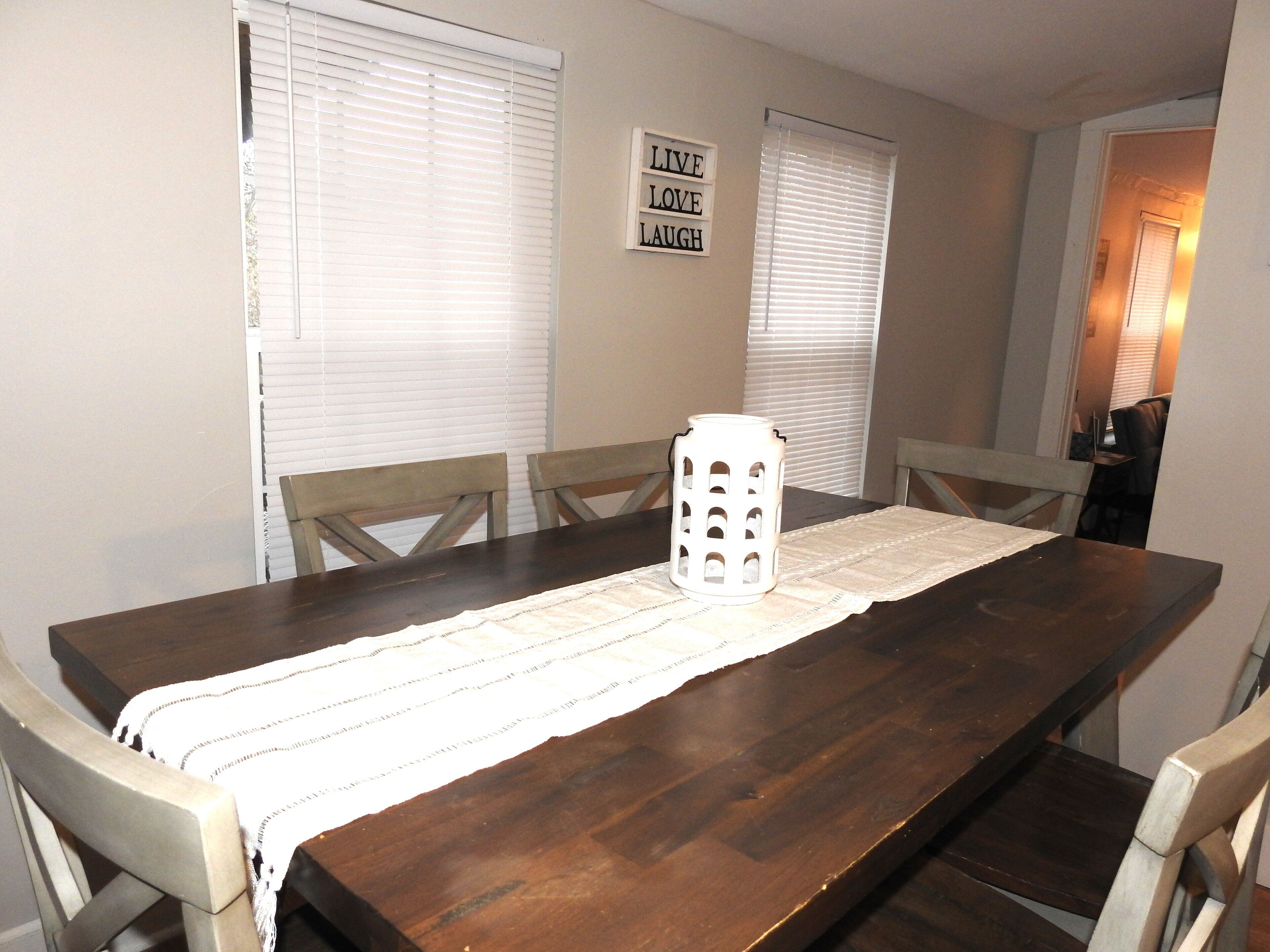 A dining table with a placemat, surrounded by chairs, in a room with two windows and a sign on the wall.