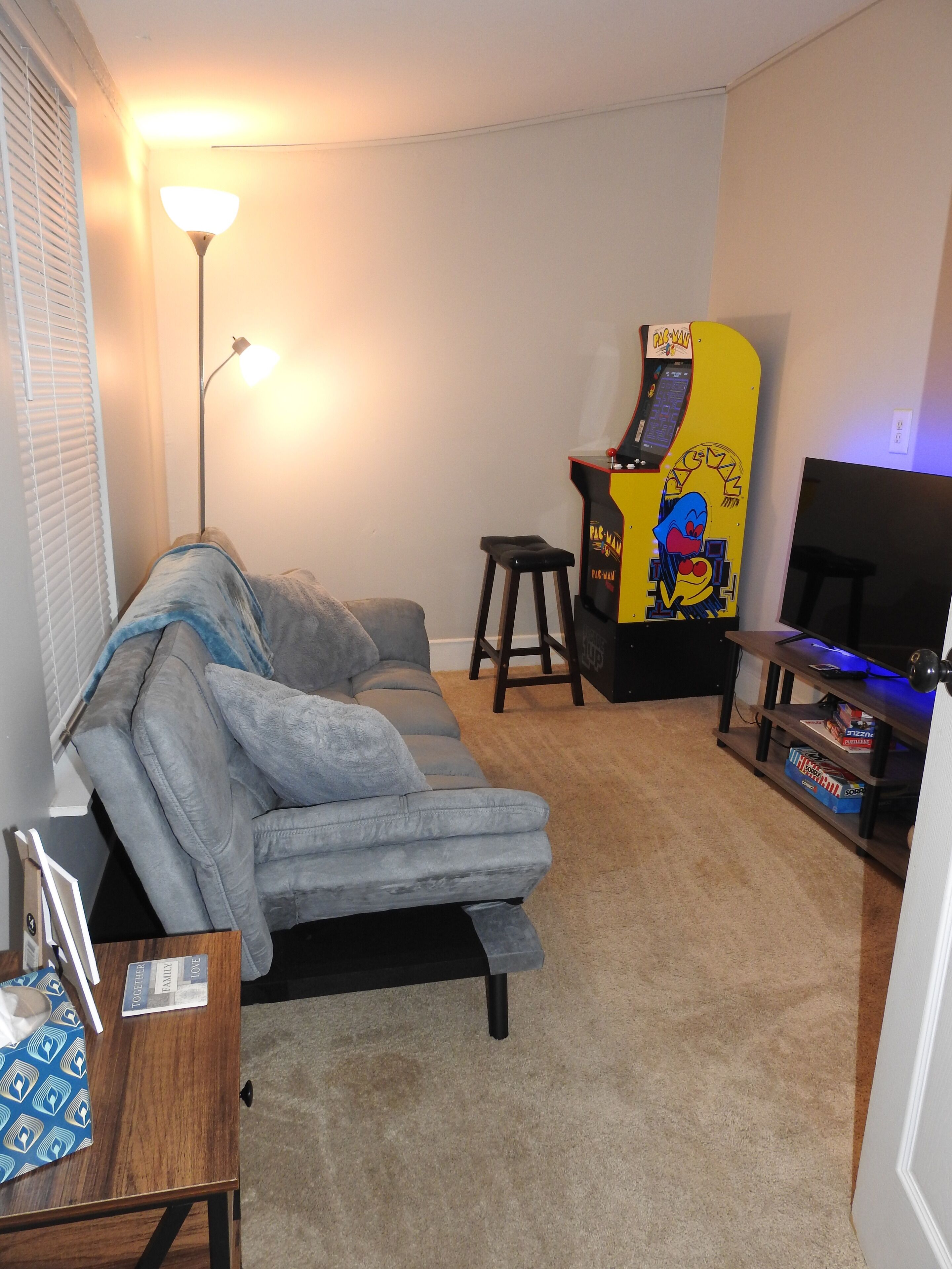 A living room with a grey sofa, a small stool, a table with a blue patterned tablecloth, and a yellow arcade machine.