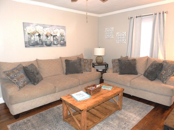 A living room with two sofas, patterned cushions, a wooden coffee table, a painting, a lamp, and a window with curtains.