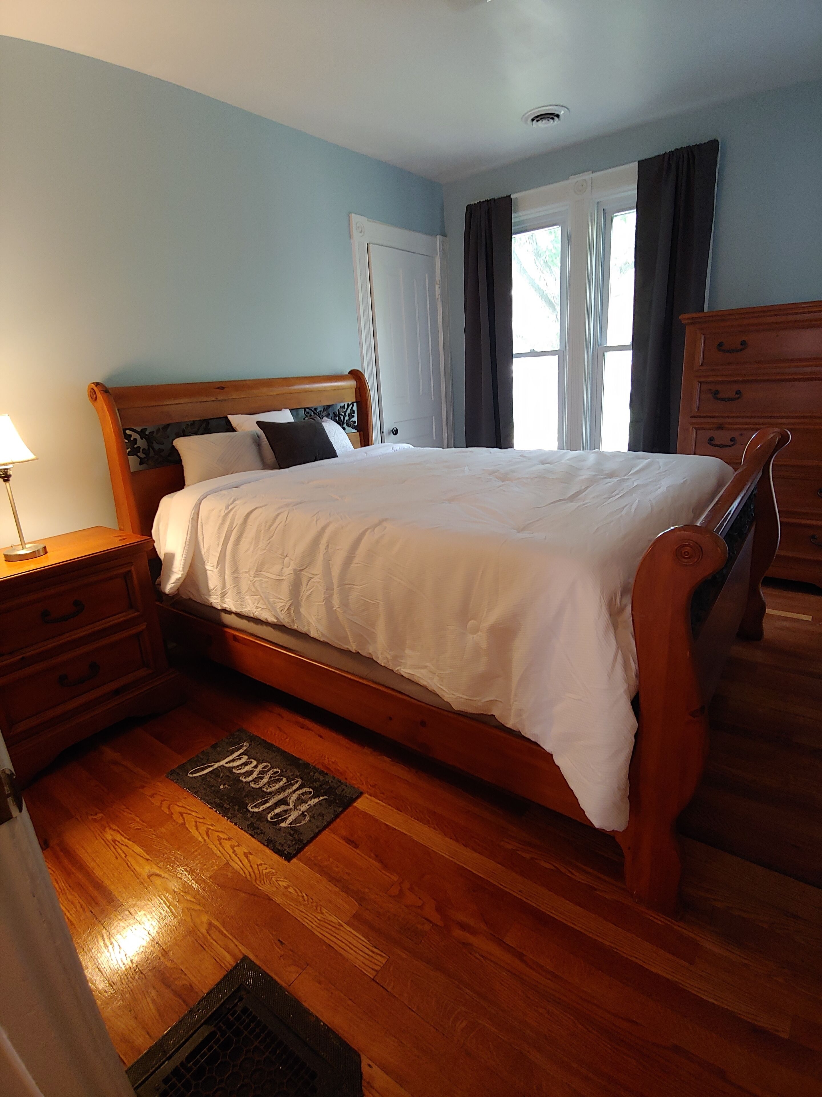 A bedroom with a wooden bed, a nightstand with a lamp, a dresser, and a window with curtains.