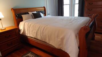 A bedroom with a wooden bed, a nightstand with a lamp, a dresser, and a window with curtains.