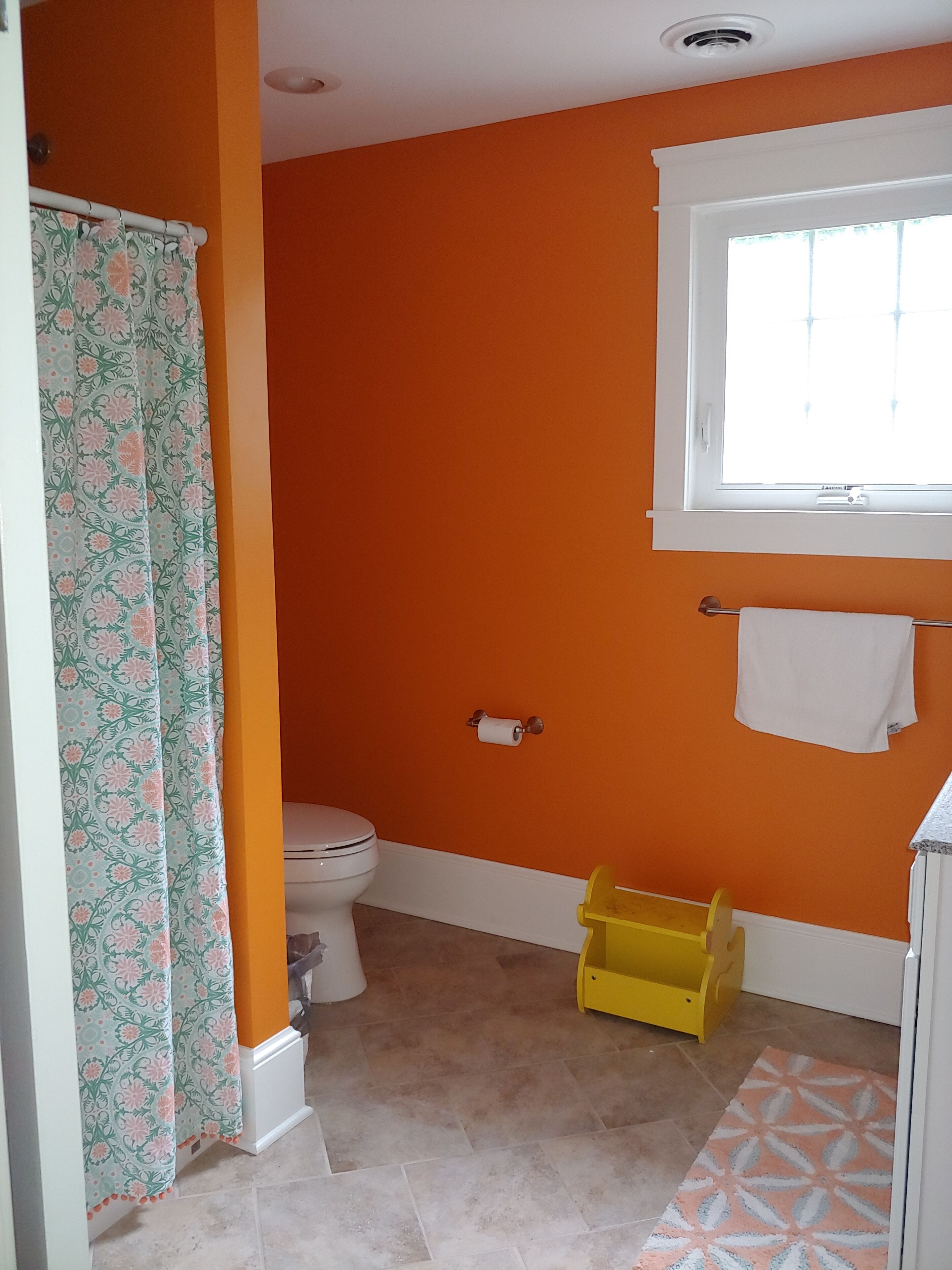 A bathroom with orange walls, a toilet, a shower curtain, a window, and a yellow storage box.